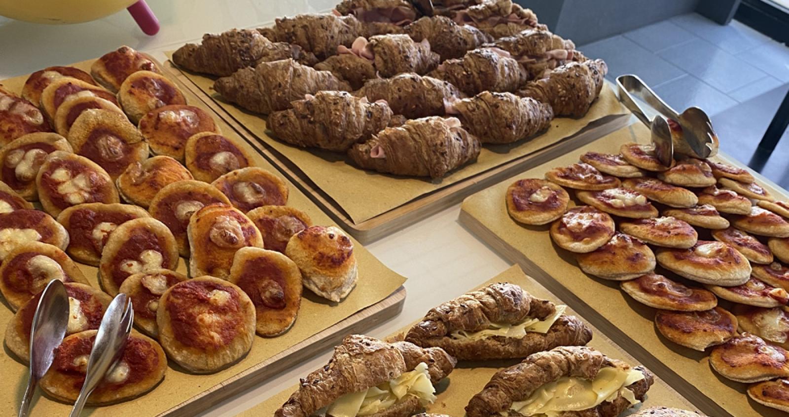 Buffet con cornetti farciti e pizzette su vassoi, posate in acciaio.