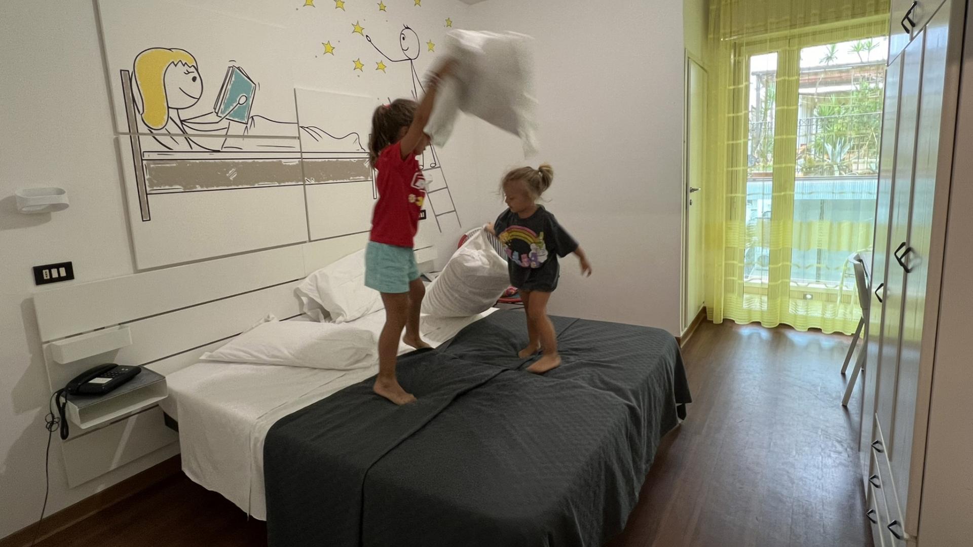 Bambini giocano con i cuscini su un letto in una stanza luminosa.