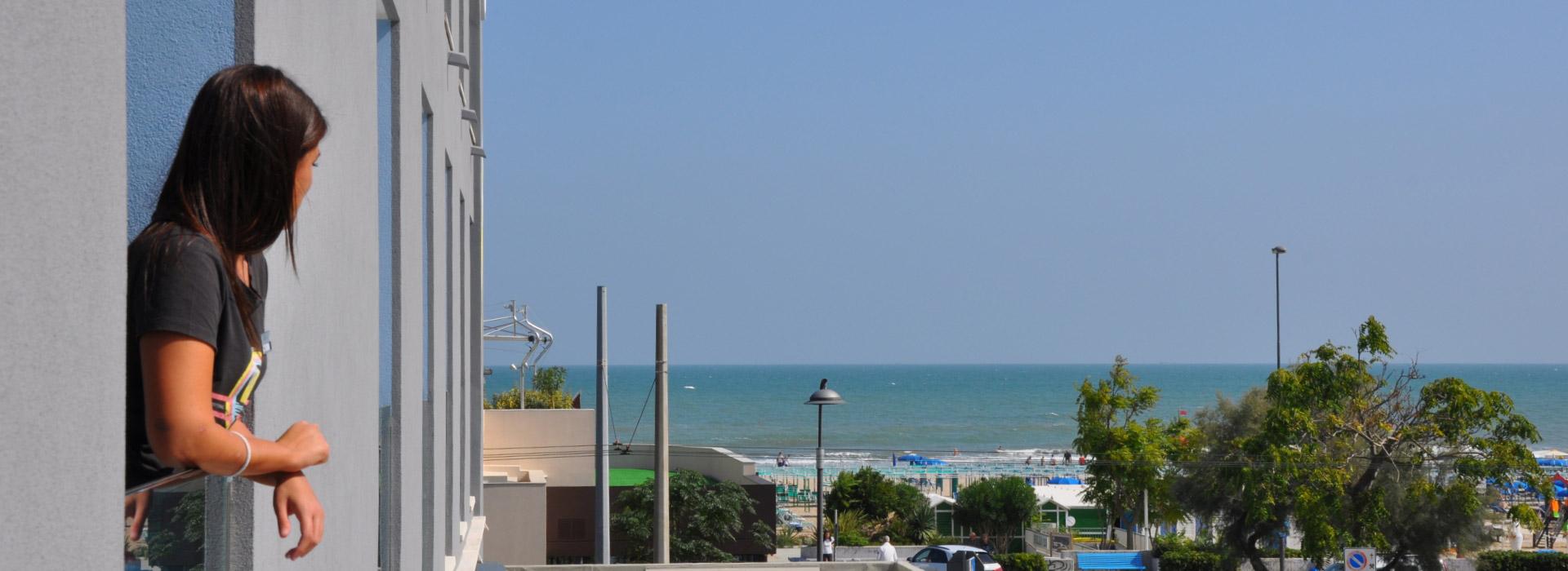 Donna guarda il mare da una finestra, giornata soleggiata e cielo sereno.