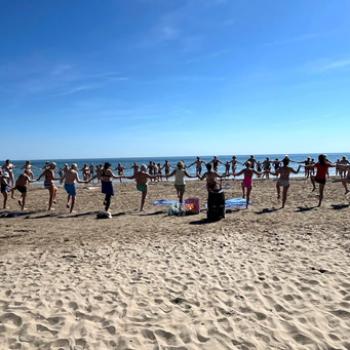 Gruppo di persone sorridenti in spiaggia attorno a un tavolo con cibo.