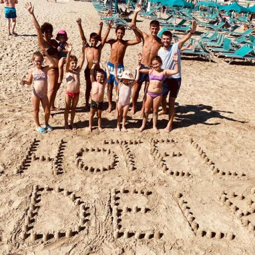 Gruppo di persone sulla spiaggia con scritta 'HOTEL ADELM' fatta di sabbia.