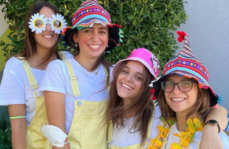 Quattro ragazze sorridenti con cappelli colorati e occhiali divertenti, indossano salopette gialle.